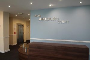 Reception area with a dark wooden desk in the foreground. The wall behind the desk has the sign "New Alexandra Court" in silver letters on a blue background. To the left, there are two elevators and beige walls. The area is well-lit with ceiling lights, perfect for showcasing investment apartments for sale by auction.