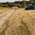 A rural landscape features a gravel path on the left and dry grass fields on the right. A blue SUV, sold by Palace Auctions, is parked in the field. The scene is marked with red lines and arrows, indicating potential paths or directions. In the background are green hills and trees.