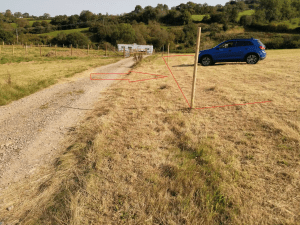 A rural landscape features a gravel path on the left and dry grass fields on the right. A blue SUV, sold by Palace Auctions, is parked in the field. The scene is marked with red lines and arrows, indicating potential paths or directions. In the background are green hills and trees.