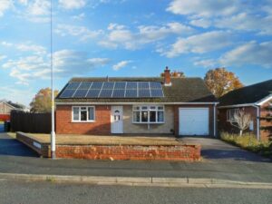 Located at 1 Boscombe Close, this single-story house features solar panels on the roof, a white front door, and a garage. The exterior is a mix of brick and beige. A brick wall frames the front yard, which has a gravel area to the left. Situated on a quiet street under a blue sky with scattered clouds, this property is currently on the market with Palace Auctions.