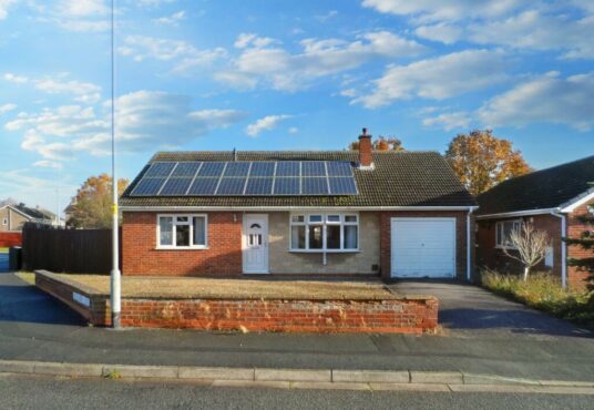 Located at 1 Boscombe Close, this single-story house features solar panels on the roof, a white front door, and a garage. The exterior is a mix of brick and beige. A brick wall frames the front yard, which has a gravel area to the left. Situated on a quiet street under a blue sky with scattered clouds, this property is currently on the market with Palace Auctions.