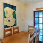 A dining room at 1 Boscombe Close, Lincoln, featuring a wooden table and four matching chairs. A large, abstract painting in white and green hues is mounted on the light-colored wall. Double glass doors with blue curtains partially reflect light, adding a subtle glow to the space. On the market with Palace Auctions.