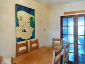 A dining room at 1 Boscombe Close, Lincoln, featuring a wooden table and four matching chairs. A large, abstract painting in white and green hues is mounted on the light-colored wall. Double glass doors with blue curtains partially reflect light, adding a subtle glow to the space. On the market with Palace Auctions.