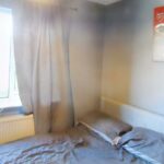 A small bedroom at 1 Boscombe Close Lincoln features grey walls and a window adorned with grey curtains. The bed, draped in matching grey bedding, is positioned against the wall. A framed poster of a football stadium hangs partially visible above the bed, with light filtering through the window. Now on the market with Palace Auctions.