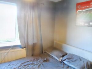 A small bedroom at 1 Boscombe Close Lincoln features grey walls and a window adorned with grey curtains. The bed, draped in matching grey bedding, is positioned against the wall. A framed poster of a football stadium hangs partially visible above the bed, with light filtering through the window. Now on the market with Palace Auctions.
