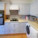 A cozy kitchen at 1 Boscombe Close Lincoln features white cabinets, a stove with a pot on it, a double oven, and various appliances including a washing machine, toaster, and slow cooker. Towels hang on the oven handle. The countertops are beige, and the wooden floors complete this charming space now on the market with Palace Auctions.