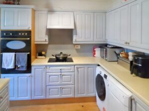 A cozy kitchen at 1 Boscombe Close Lincoln features white cabinets, a stove with a pot on it, a double oven, and various appliances including a washing machine, toaster, and slow cooker. Towels hang on the oven handle. The countertops are beige, and the wooden floors complete this charming space now on the market with Palace Auctions.
