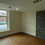 A minimalistic room with wooden flooring, beige walls, a large window on the left, and a wall on the right featuring dark stacked stone cladding with a rectangular recessed niche. The window shows a partial view of a red brick building outside.