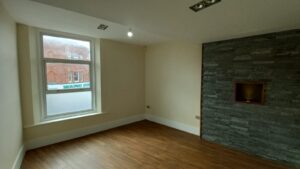 A minimalistic room with wooden flooring, beige walls, a large window on the left, and a wall on the right featuring dark stacked stone cladding with a rectangular recessed niche. The window shows a partial view of a red brick building outside.