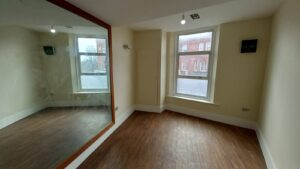 A small, empty room with a large mirror on the left wall, two large windows letting in natural light on the right, laminate wooden flooring, and cream-colored walls. A single small light bulb hangs from the white ceiling.
