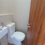 A small, modern bathroom featuring a white toilet and a compact white sink with a silver faucet. The walls are tiled in a light, neutral color and the floor has wood-patterned tiles. A wooden door with a silver handle is partially open.