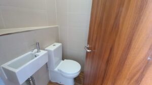 A small, modern bathroom featuring a white toilet and a compact white sink with a silver faucet. The walls are tiled in a light, neutral color and the floor has wood-patterned tiles. A wooden door with a silver handle is partially open.