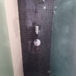 A modern shower area at 120 St Margaret's Road Twickenham boasts dark square tiles, a wall-mounted shower head, a soap dispenser, and a metallic faucet handle. Frosted glass panels on either side enhance its sleek, minimalistic look.