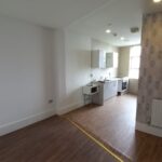 A modern, unfurnished kitchen and living space at 120 St Margaret's Road Twickenham features wooden flooring. The kitchen area boasts white cabinets, a stove, and an oven. One living area wall is white, while another showcases patterned wallpaper. A yellow tape line neatly separates the two spaces.