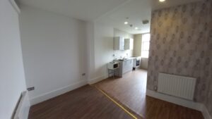 A modern, unfurnished kitchen and living space at 120 St Margaret's Road Twickenham features wooden flooring. The kitchen area boasts white cabinets, a stove, and an oven. One living area wall is white, while another showcases patterned wallpaper. A yellow tape line neatly separates the two spaces.