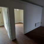 A view of two open doorways in an empty apartment at 120 St Margaret's Road, Twickenham, with hardwood floors. The room on the left has a window letting in natural light, while the room on the right is dimly lit. There is a small heater installed on the wall in the foreground.