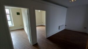 A view of two open doorways in an empty apartment at 120 St Margaret's Road, Twickenham, with hardwood floors. The room on the left has a window letting in natural light, while the room on the right is dimly lit. There is a small heater installed on the wall in the foreground.