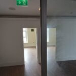 A sparsely furnished room with white walls and wooden flooring. The room has large windows allowing natural light in. There's a doorway in the center, leading to another similar room. A green emergency exit sign is mounted on the ceiling above the doorway at 120 St Margaret's Road Twickenham.