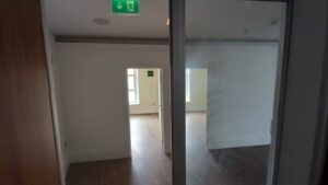 A sparsely furnished room with white walls and wooden flooring. The room has large windows allowing natural light in. There's a doorway in the center, leading to another similar room. A green emergency exit sign is mounted on the ceiling above the doorway at 120 St Margaret's Road Twickenham.