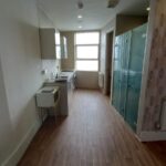 Situated at 120 St Margaret's Road Twickenham, this narrow kitchen boasts wooden flooring, white walls, and a large window at the end. It features a small standing microwave, a sink, and a compact stove on the left side. The right wall has patterned wallpaper with a radiator below it.