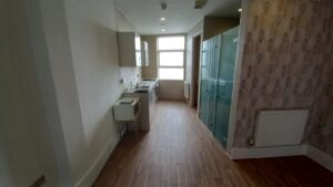 Situated at 120 St Margaret's Road Twickenham, this narrow kitchen boasts wooden flooring, white walls, and a large window at the end. It features a small standing microwave, a sink, and a compact stove on the left side. The right wall has patterned wallpaper with a radiator below it.
