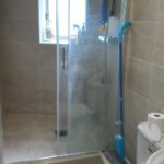 A bathroom at 120 St Margaret's Road Twickenham features beige-tiled walls and floor, a glass-enclosed shower, a white toilet with a roll of toilet paper on top, and a mop leaning against the wall. The sink has a dark pump bottle beside it, while the window allows natural light to enter.