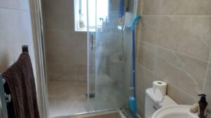 A bathroom at 120 St Margaret's Road Twickenham features beige-tiled walls and floor, a glass-enclosed shower, a white toilet with a roll of toilet paper on top, and a mop leaning against the wall. The sink has a dark pump bottle beside it, while the window allows natural light to enter.