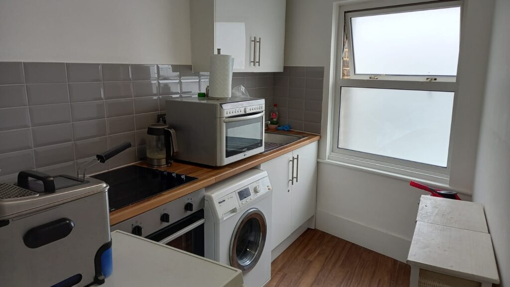 A compact kitchen with white cabinets, a wooden countertop, and gray tiled backsplash. Appliances include a washing machine, an electric stovetop with an oven, a toaster, and a kettle. A window with frosted glass is on the right side, and there is a small table below it.