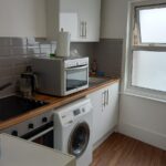 A compact kitchen with white cabinets, a wooden countertop, and gray tiled backsplash. Appliances include a washing machine, an electric stovetop with an oven, a toaster, and a kettle. A window with frosted glass is on the right side, and there is a small table below it.