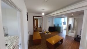 A modern living area with wooden floors featuring a wall-mounted TV, wooden coffee tables, and a mirror on the wall. To the left is a partially visible bedroom with a bed. The kitchen with white cabinets and appliances is visible in the background.