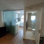 A small modern apartment features a living area with a flat-screen TV on the left, a kitchen with white cabinets and a counter in the middle, and a bathroom with a toilet and shower on the right. The space is lit by natural light from windows and overhead lighting.