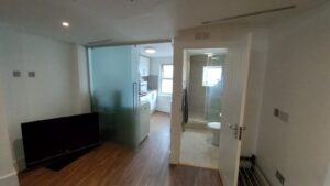 A small modern apartment features a living area with a flat-screen TV on the left, a kitchen with white cabinets and a counter in the middle, and a bathroom with a toilet and shower on the right. The space is lit by natural light from windows and overhead lighting.