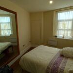 A small, tidy bedroom with a single bed against the wall. There is a large mirror on the left wall, reflecting the bed and showing a window with sheer white curtains. The bed has white bedding with purple accents. The room is painted in light colors.