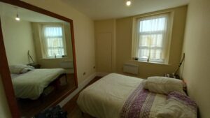 A small, tidy bedroom with a single bed against the wall. There is a large mirror on the left wall, reflecting the bed and showing a window with sheer white curtains. The bed has white bedding with purple accents. The room is painted in light colors.