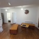 A modern room with wooden flooring, featuring a large mirror on one wall, a wooden table, and a small white foldable table with a laptop. A collage picture frame hangs on the wall. A door leads to another room with a bed visible. The room is well-lit with ceiling lights.