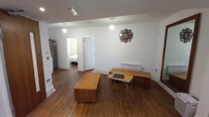 A modern room with wooden flooring, featuring a large mirror on one wall, a wooden table, and a small white foldable table with a laptop. A collage picture frame hangs on the wall. A door leads to another room with a bed visible. The room is well-lit with ceiling lights.