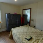 A small bedroom with a double bed covered in a beige quilt. The room features a window with a small portrait and decor on the sill, two wardrobe-like storage units, a floor lamp, and visible wooden flooring. The door to an adjacent room is open.