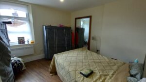 A small bedroom with a double bed covered in a beige quilt. The room features a window with a small portrait and decor on the sill, two wardrobe-like storage units, a floor lamp, and visible wooden flooring. The door to an adjacent room is open.