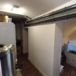 A narrow room with a slanted ceiling, featuring a large metal container on the left, possibly a water heater or boiler. The floor is wooden, and the room contains various boxes and items, including cabinets and shelves. The lighting is provided by wall-mounted fixtures.