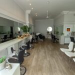 A brightly lit, modern hair salon located at 120 St Margaret's Road Twickenham features white walls and wooden flooring. It boasts multiple styling stations with large mirrors, black chairs, and white waiting area chairs. Plants and minimalistic decor add a touch of greenery and elegance.
