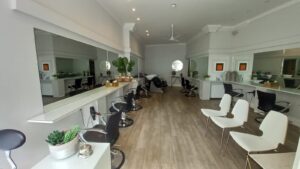 A brightly lit, modern hair salon located at 120 St Margaret's Road Twickenham features white walls and wooden flooring. It boasts multiple styling stations with large mirrors, black chairs, and white waiting area chairs. Plants and minimalistic decor add a touch of greenery and elegance.
