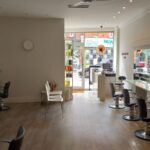 A modern hair salon at 120 St Margaret's Road Twickenham features multiple styling stations, each equipped with mirrors and chairs. The room boasts sleek, white decor with plants placed around for a fresh look. A large window and glass door at the front let in natural light, offering a street view outside.
