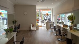 A modern hair salon at 120 St Margaret's Road Twickenham features multiple styling stations, each equipped with mirrors and chairs. The room boasts sleek, white decor with plants placed around for a fresh look. A large window and glass door at the front let in natural light, offering a street view outside.