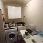 A small, narrow kitchen with white cabinets, countertops, and walls. A washing machine is installed under the counter. The counter is cluttered with various kitchen items. A table with a plate, cup, and containers is placed in the middle of the room.