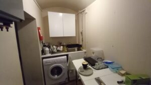 A small, narrow kitchen with white cabinets, countertops, and walls. A washing machine is installed under the counter. The counter is cluttered with various kitchen items. A table with a plate, cup, and containers is placed in the middle of the room.