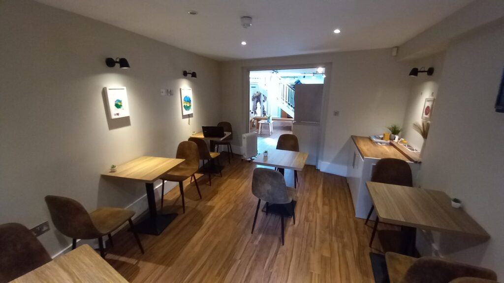 A small café interior with wooden floors, beige walls, and several square wooden tables with brown chairs. The walls have minimalist artwork. The room is well-lit with ceiling lights, and a doorway at the back leads to a brighter area with a staircase.