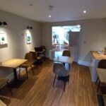 A small café interior with wooden floors, beige walls, and several square wooden tables with brown chairs. The walls have minimalist artwork. The room is well-lit with ceiling lights, and a doorway at the back leads to a brighter area with a staircase.