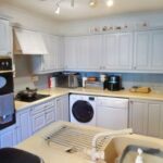 A clean, modern kitchen at 1 Boscombe Close Lincoln, featuring light-colored cabinets, a double oven, dishwasher, microwave, and washing machine. The countertops are clutter-free with a drying rack over the sink and small appliances like a toaster and coffee machine. On the market with Palace Auctions.