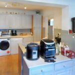 A modern kitchen at 1 Boscombe Close, Lincoln, features white cabinets, wooden flooring, and various appliances. An oven, washing machine, and dryer are tucked under the counter. Countertops hold an air fryer, rice cooker, condiments, and a soda pack. A semi-open doorway leads to an adjacent room. On the market with Palace Auctions.
