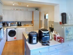 A modern kitchen at 1 Boscombe Close, Lincoln, features white cabinets, wooden flooring, and various appliances. An oven, washing machine, and dryer are tucked under the counter. Countertops hold an air fryer, rice cooker, condiments, and a soda pack. A semi-open doorway leads to an adjacent room. On the market with Palace Auctions.